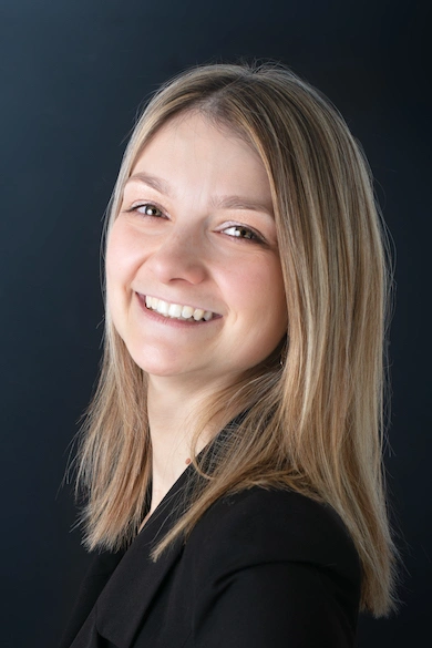 Elodie_Defosse Portrait d’une femme souriante aux cheveux blonds raides, portant une veste noire, posant de profil devant un fond sombre.